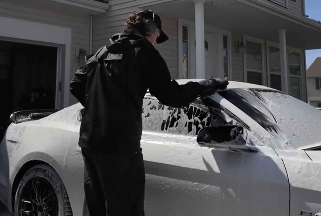 A Waypoint Auto Detailing technician hand-washing a white sports car covered in thick foam suds in a residential driveway in Spokane Valley.
