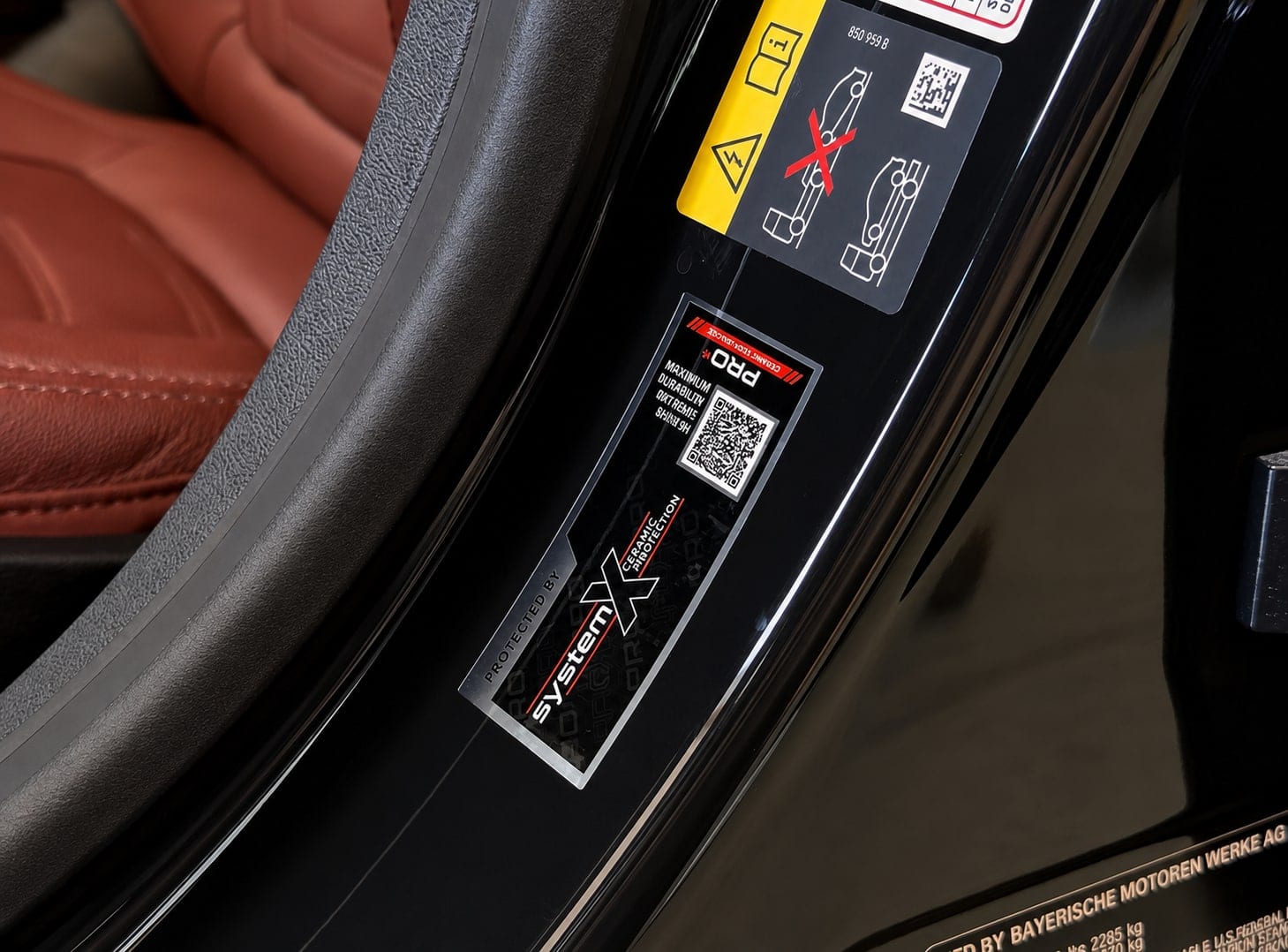 Close-up of a car door jamb showing a black BMW high-voltage warning label alongside a red and black 'System X Ceramic Protection PRO' sticker, with a red leather seat visible in the background.