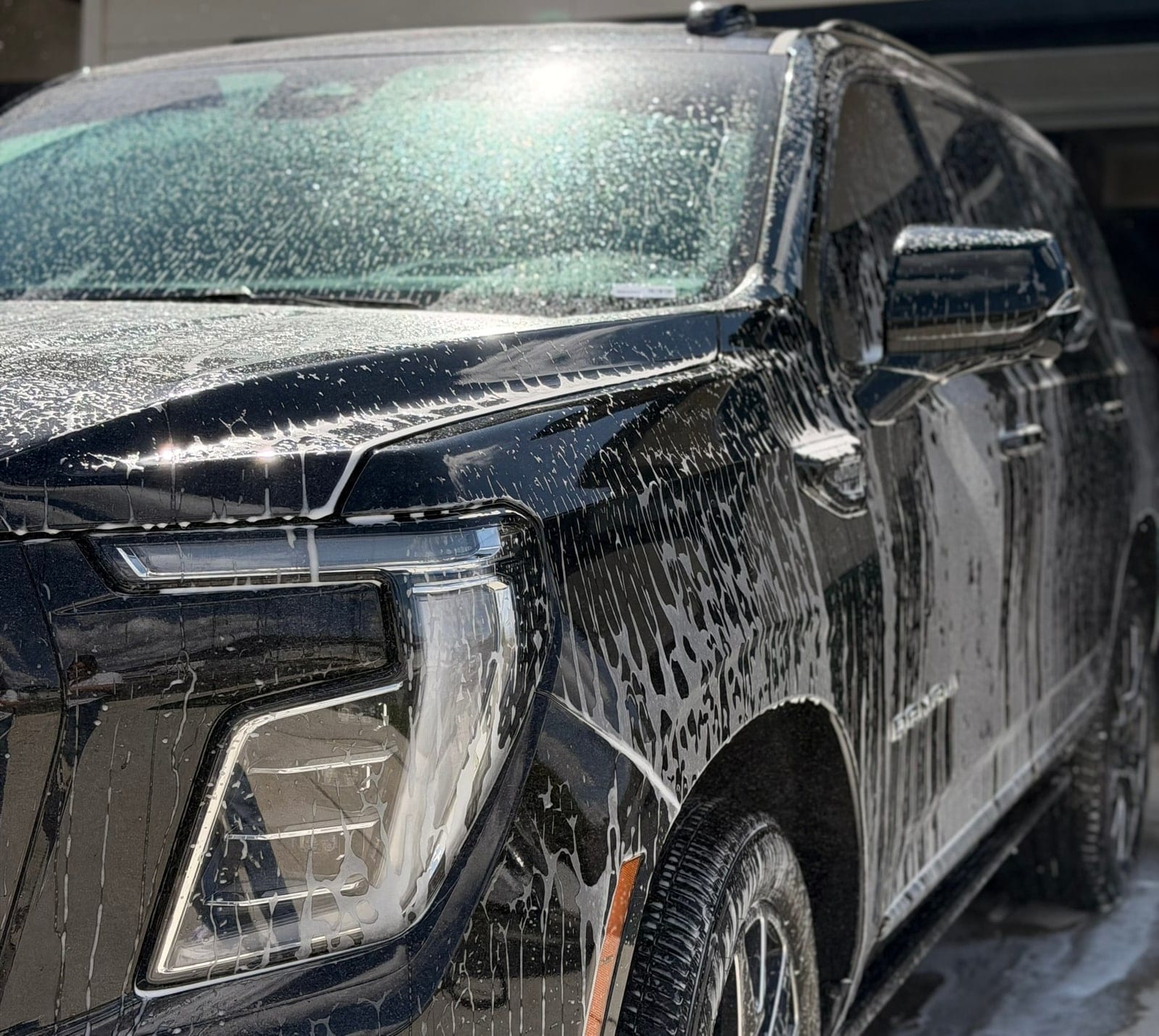 A black SUV, badged 'Denali', is covered in thick foam soap during an exterior hand wash, with close focus on the front headlight, hood, and fender.