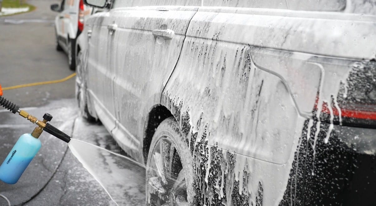 A dark SUV covered in thick white foam is being pre-washed with a snow foam cannon attached to a pressure washer in a parking lot.