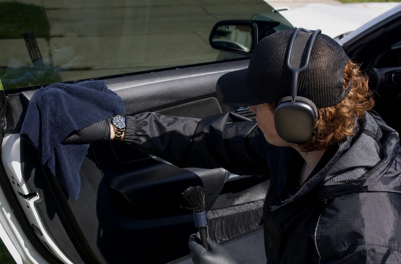A detailer wearing a cap, headphones, and gloves reaches into a white car's interior with a blue microfiber towel, holding a detailing brush, wiping down the dashboard and door panel.