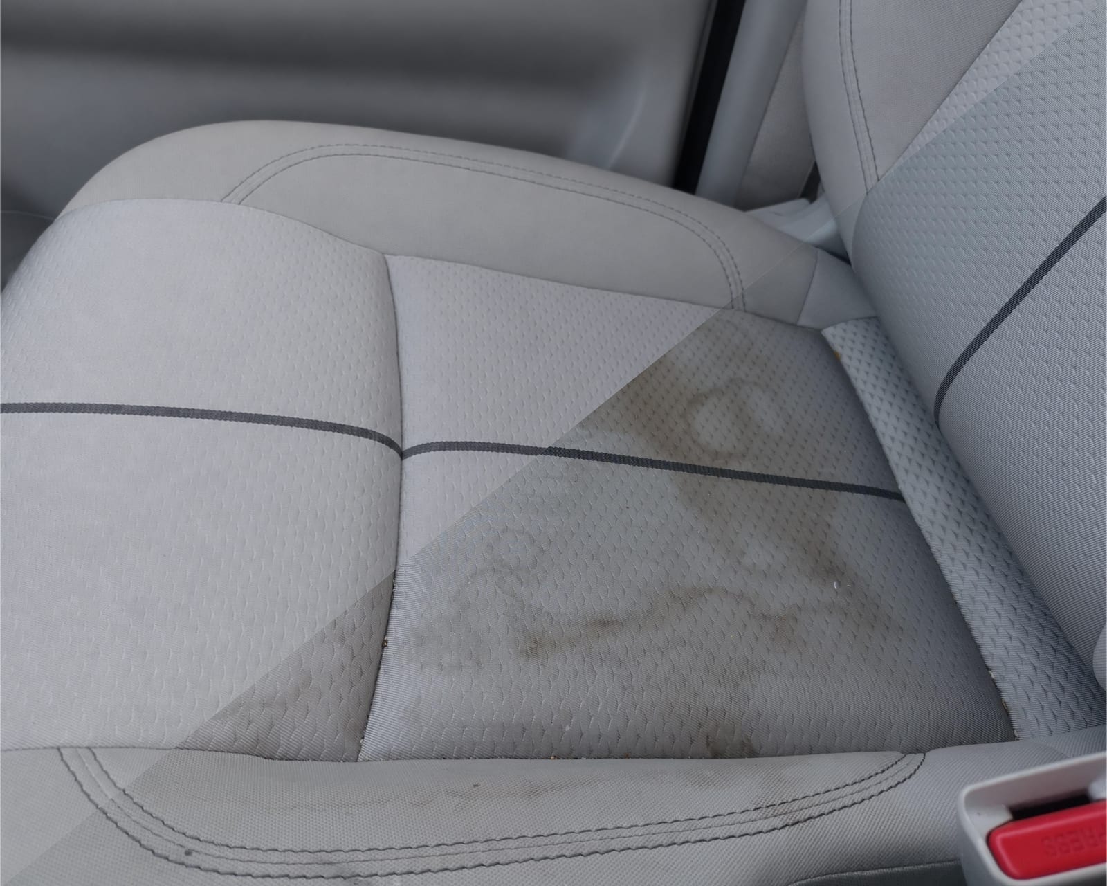 A gray fabric car seat with a diagonal split showing a visible coffee stain on one half and clean, spot-free fabric on the other half.