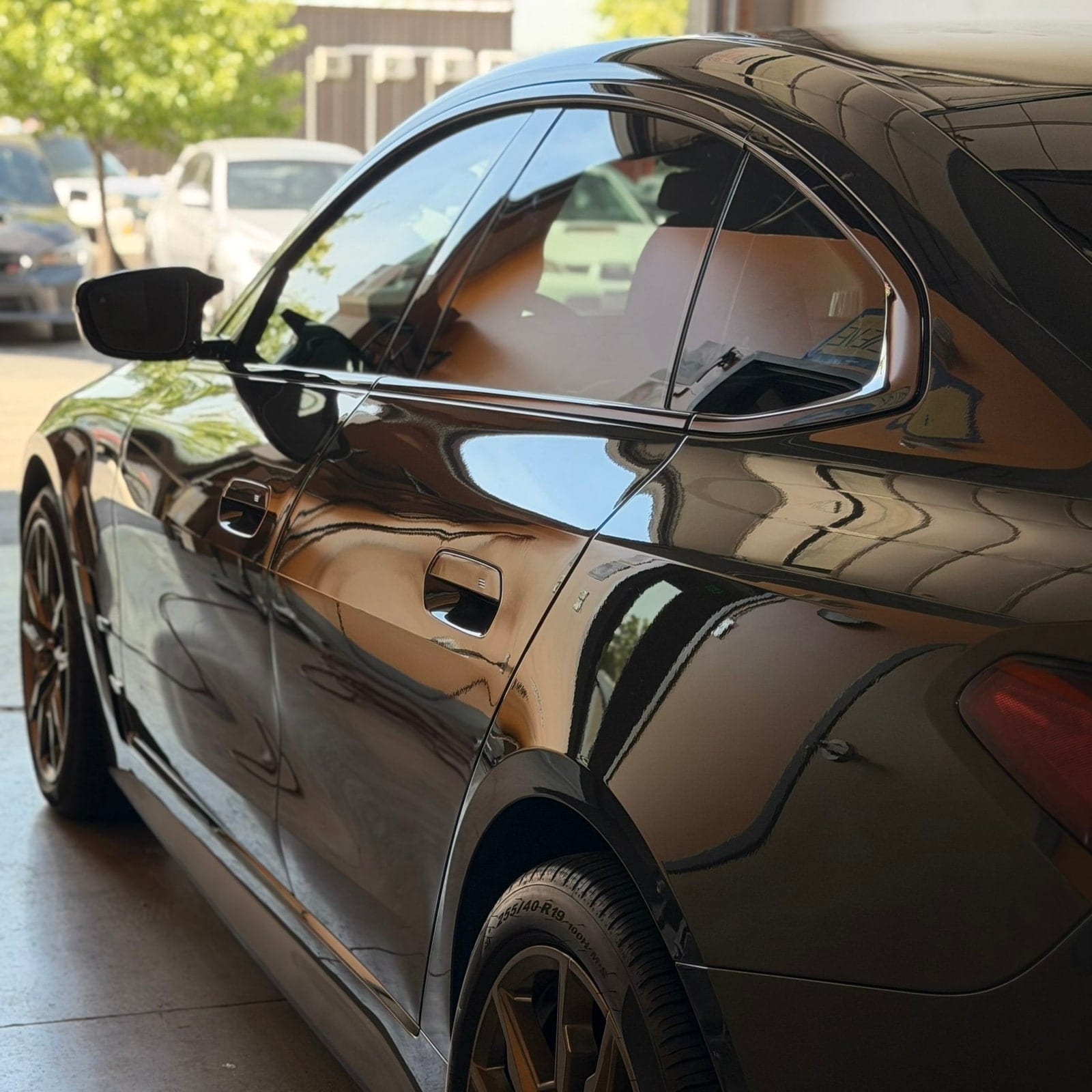 Close-up side profile of a black coupe with a highly glossy, reflective paint finish parked in a garage, showing sharp reflections of surrounding vehicles and trees.