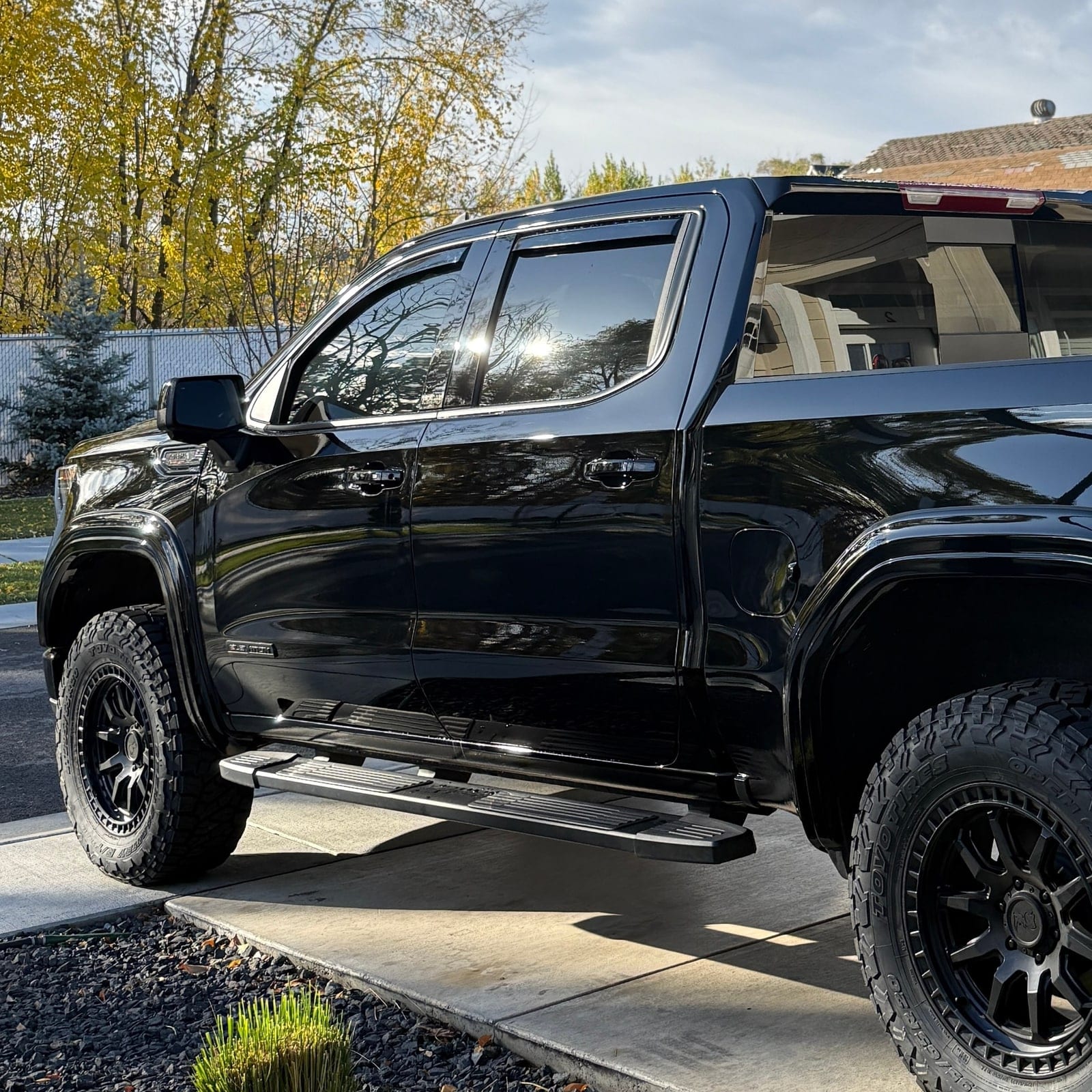 A black lifted pickup truck with aftermarket off-road wheels and tires is parked in a driveway, its exterior paint and windows showing a glossy, clean finish.