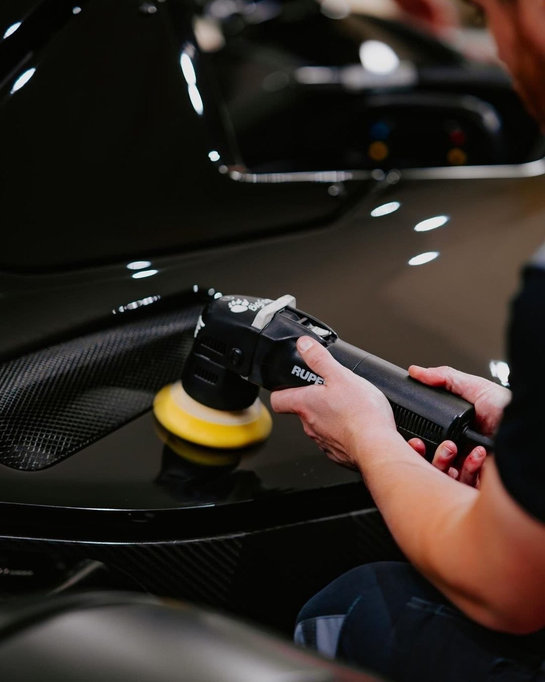 A person uses a black dual-action polisher with a yellow pad to buff the glossy black paint and carbon fiber trim of a car's hood/fender area.