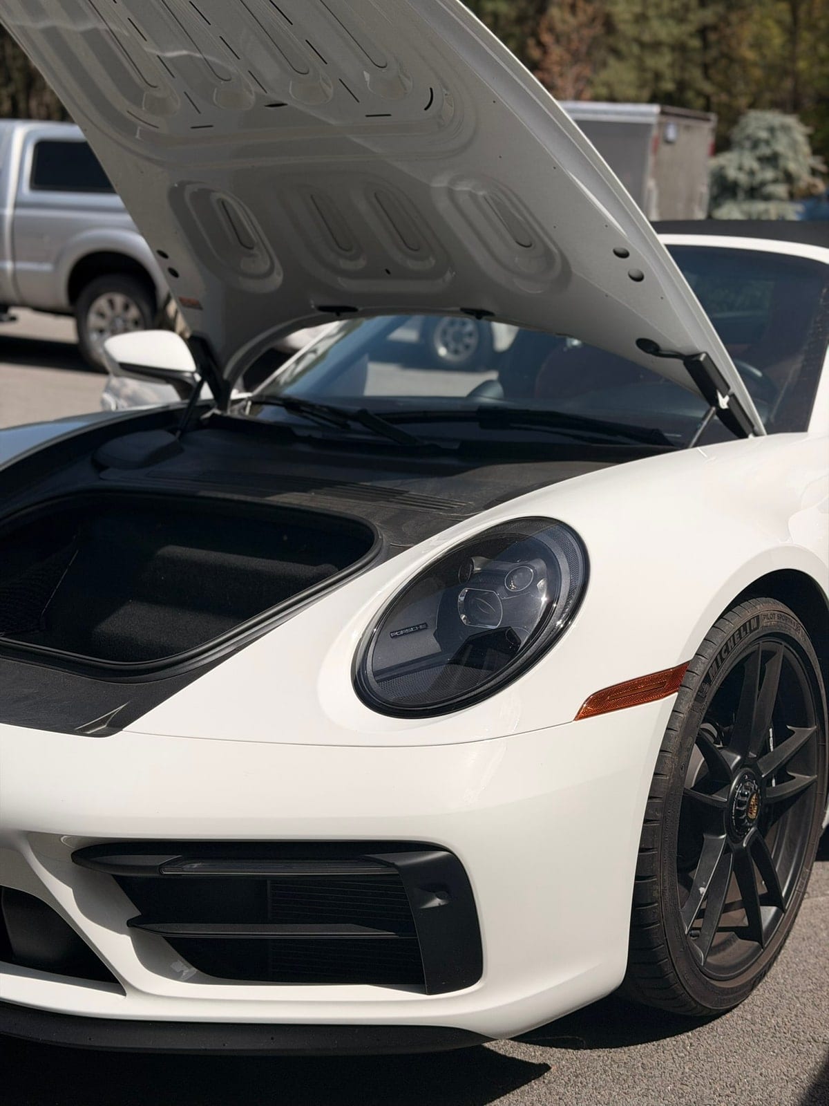 A white Porsche 911 sports car parked outdoors with its front trunk (frunk) open, showing a close-up of the front fender, headlight, black alloy wheel, and clean paintwork.