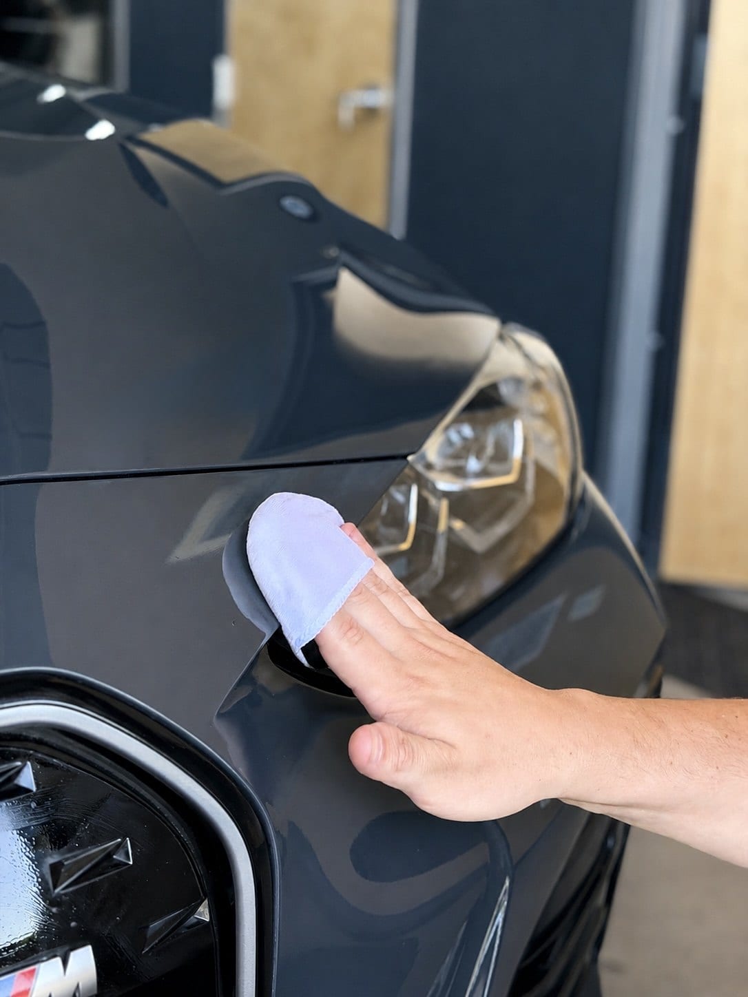 A hand uses a microfiber cloth to wipe and buff the front bumper and headlight area of a dark gray BMW M car in a garage setting.