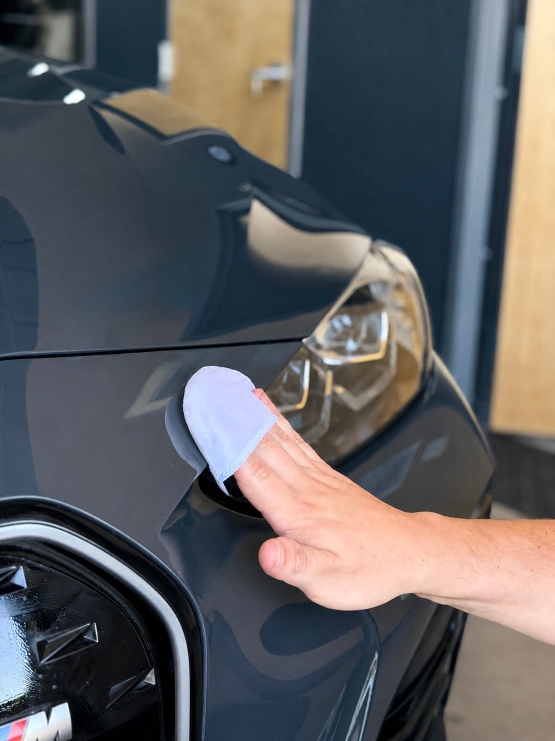A hand uses a microfiber cloth to wipe and buff the front bumper and headlight area of a dark gray BMW M car in a garage setting.