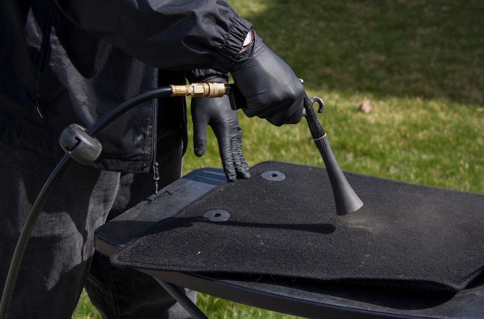 A gloved detailer uses a compressed-air blow gun to clean deep dust and debris from a black textured car floor mat, with green grass visible in the background.