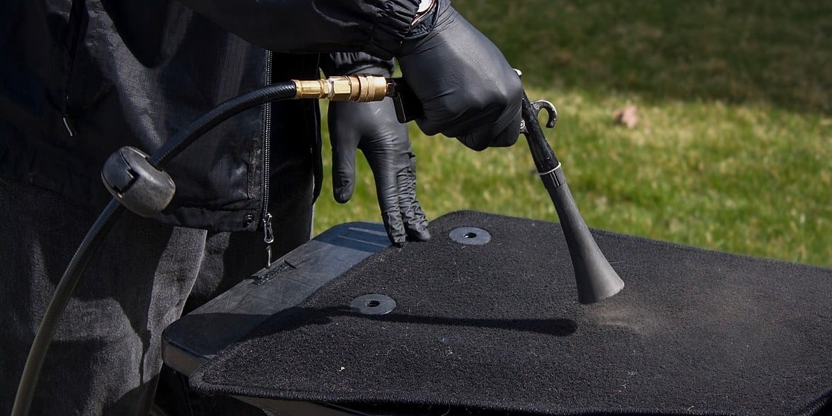 A gloved detailer uses a compressed-air blow gun to clean deep dust and debris from a black textured car floor mat, with green grass visible in the background.