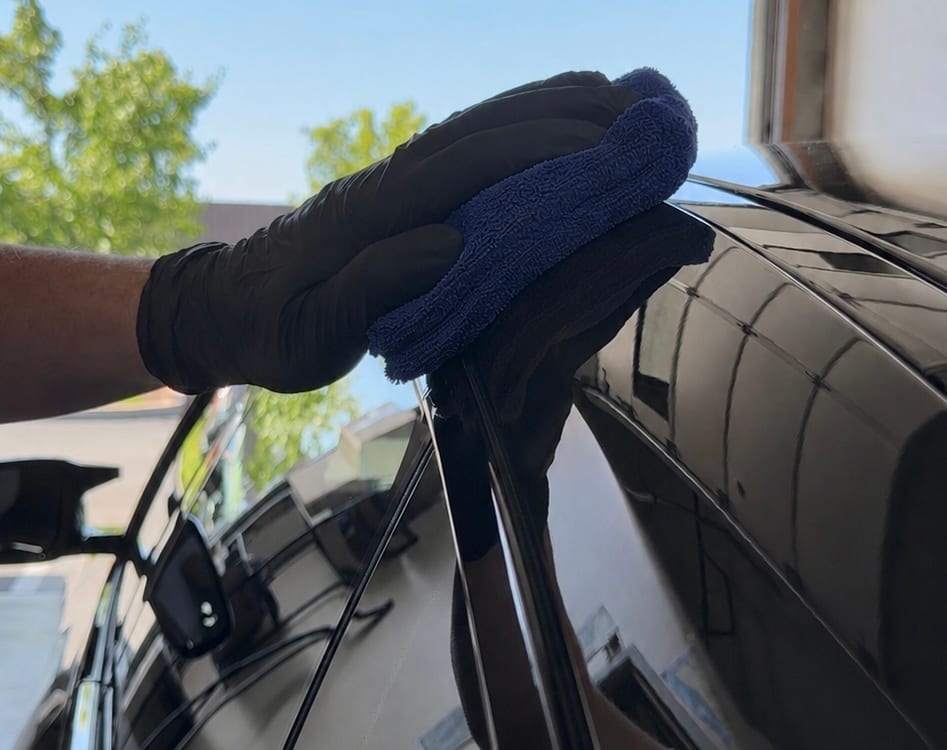 A gloved hand uses a blue microfiber applicator to spread ceramic coating onto the glossy black roofline of a vehicle, with trees reflected in the paint.