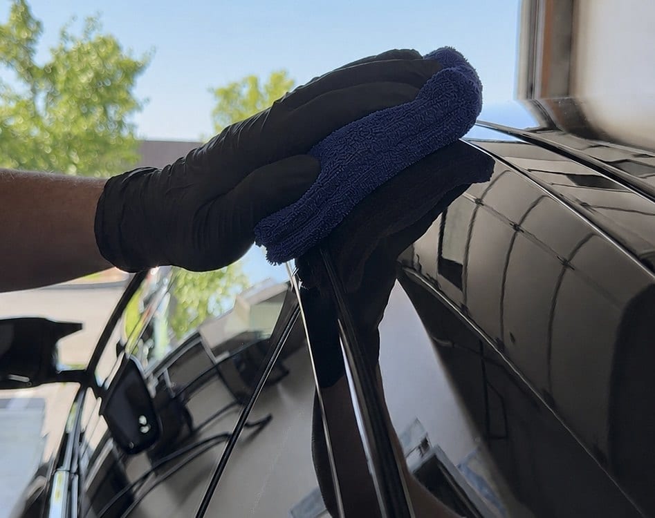 A gloved hand uses a blue microfiber applicator to wipe a coating or wax onto the glossy black exterior panel of a vehicle, with trees and sky visible in the reflection.