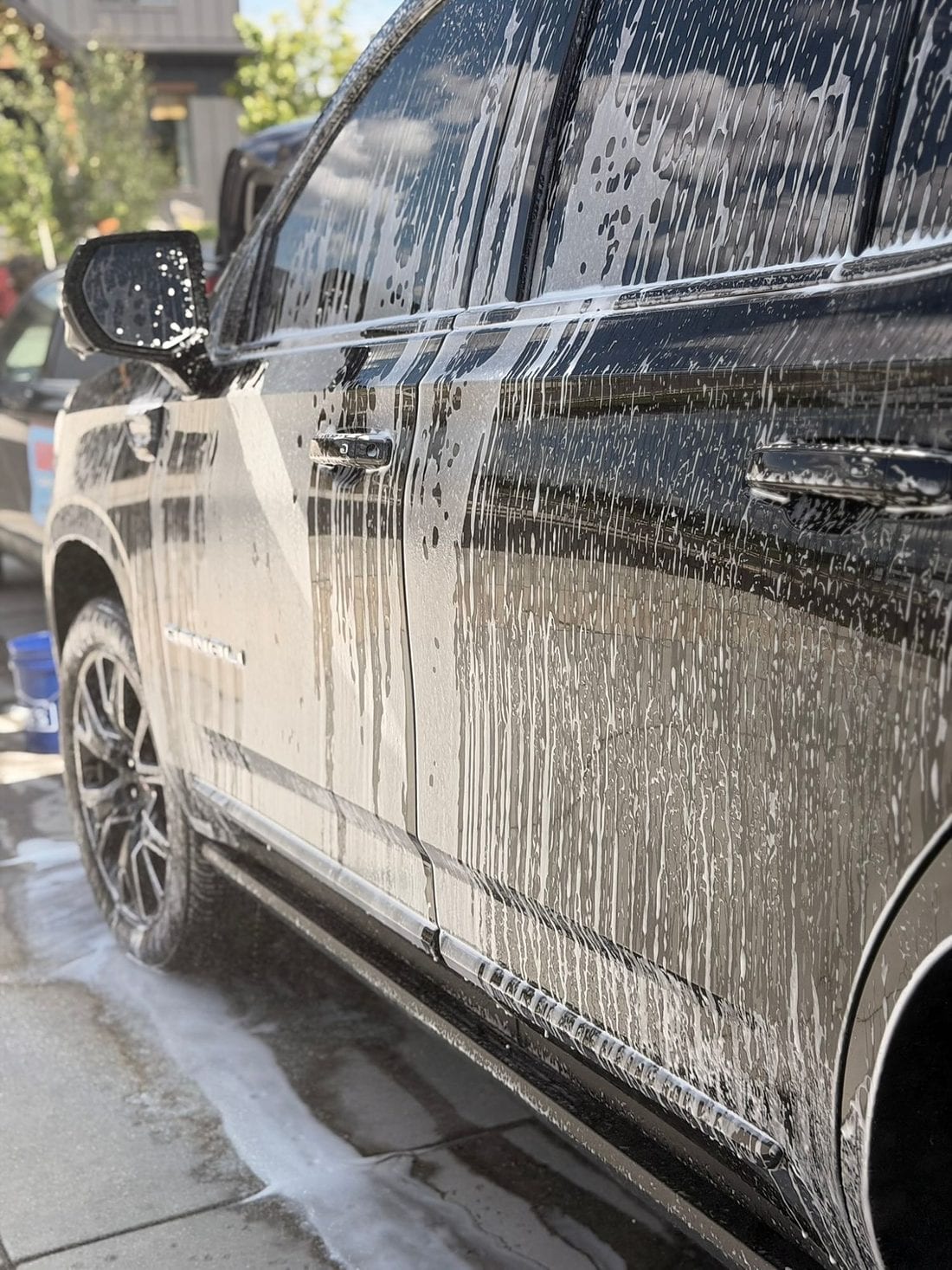 A dark GMC Denali SUV covered in thick foam soap during an exterior wash, with a blue detailing bucket visible near the front wheel.