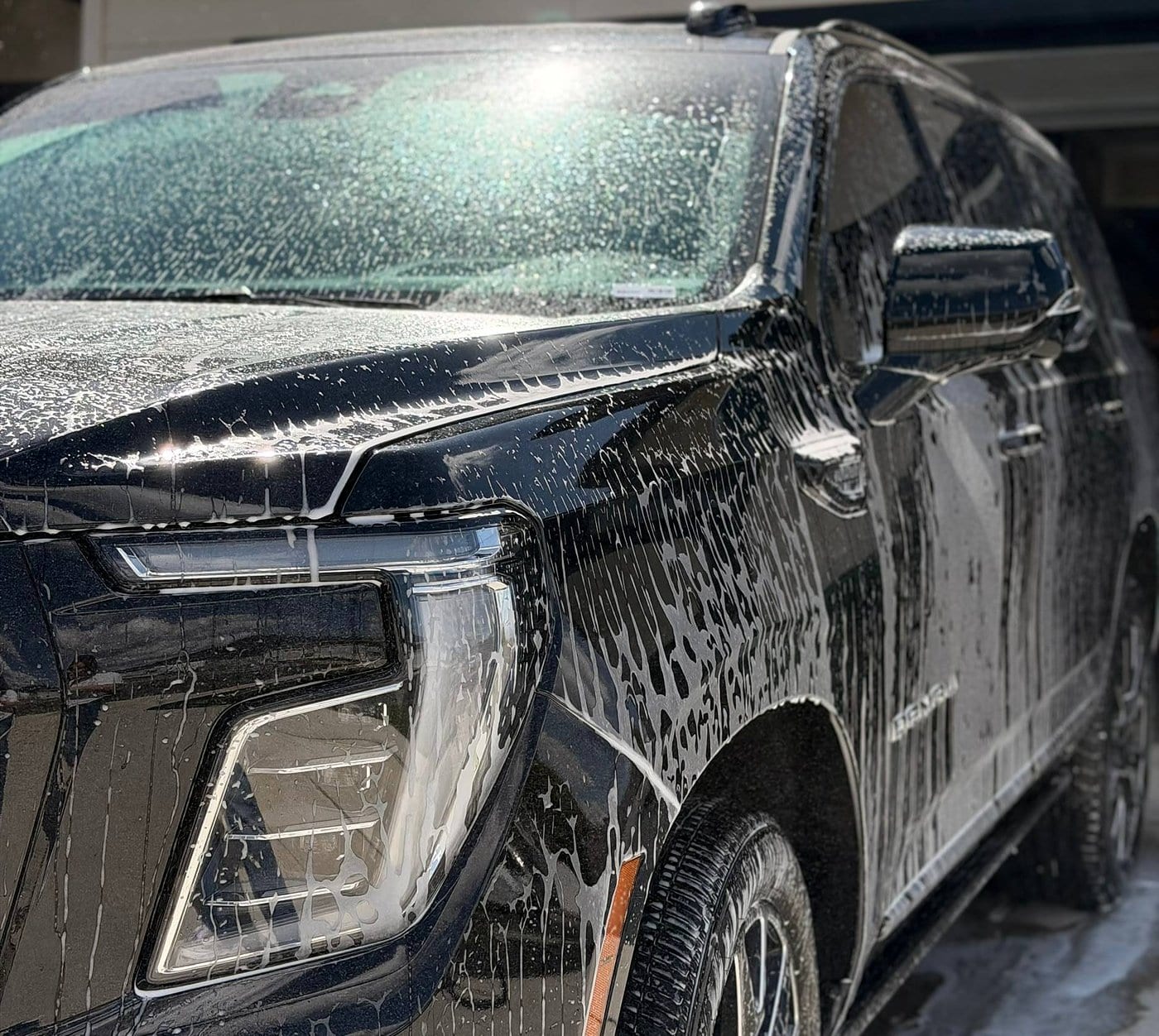 A black SUV, badged “Denali”, is covered in thick foam soap during an exterior hand wash, with close focus on the front headlight, hood, and fender.