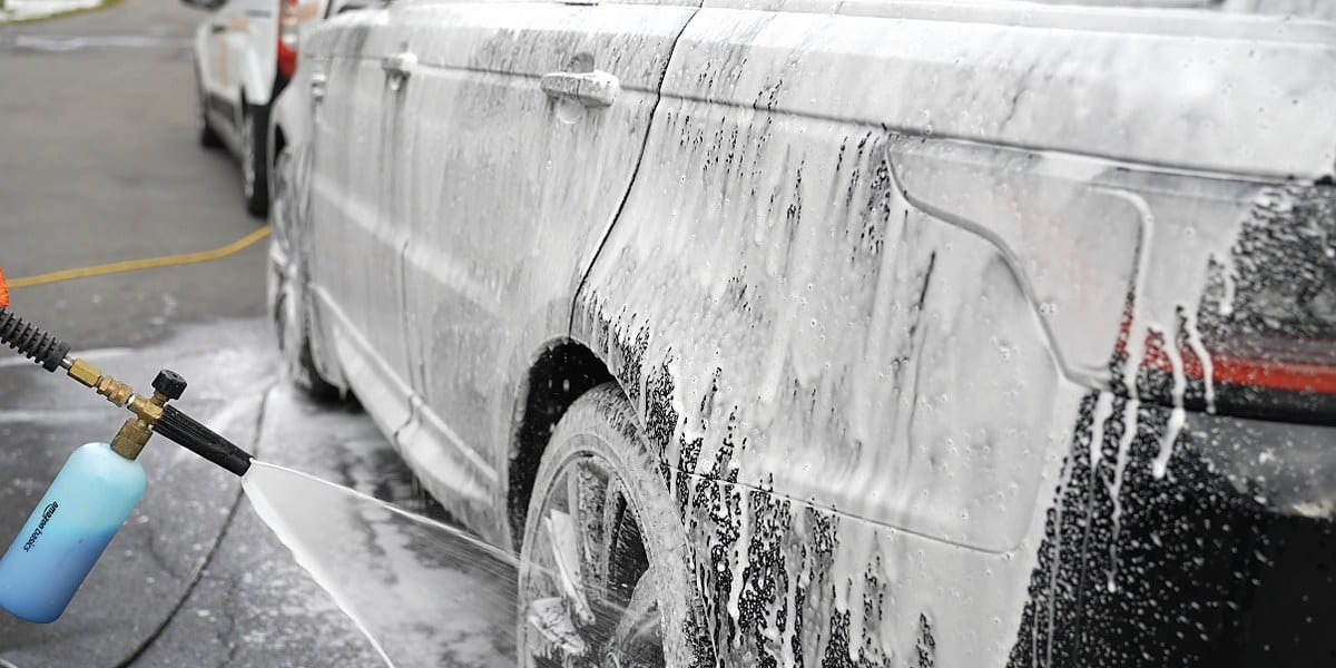 A white SUV is coated in thick white pre-wash foam from a snow-foam cannon, with foam dripping down the door panels and rear wheel.