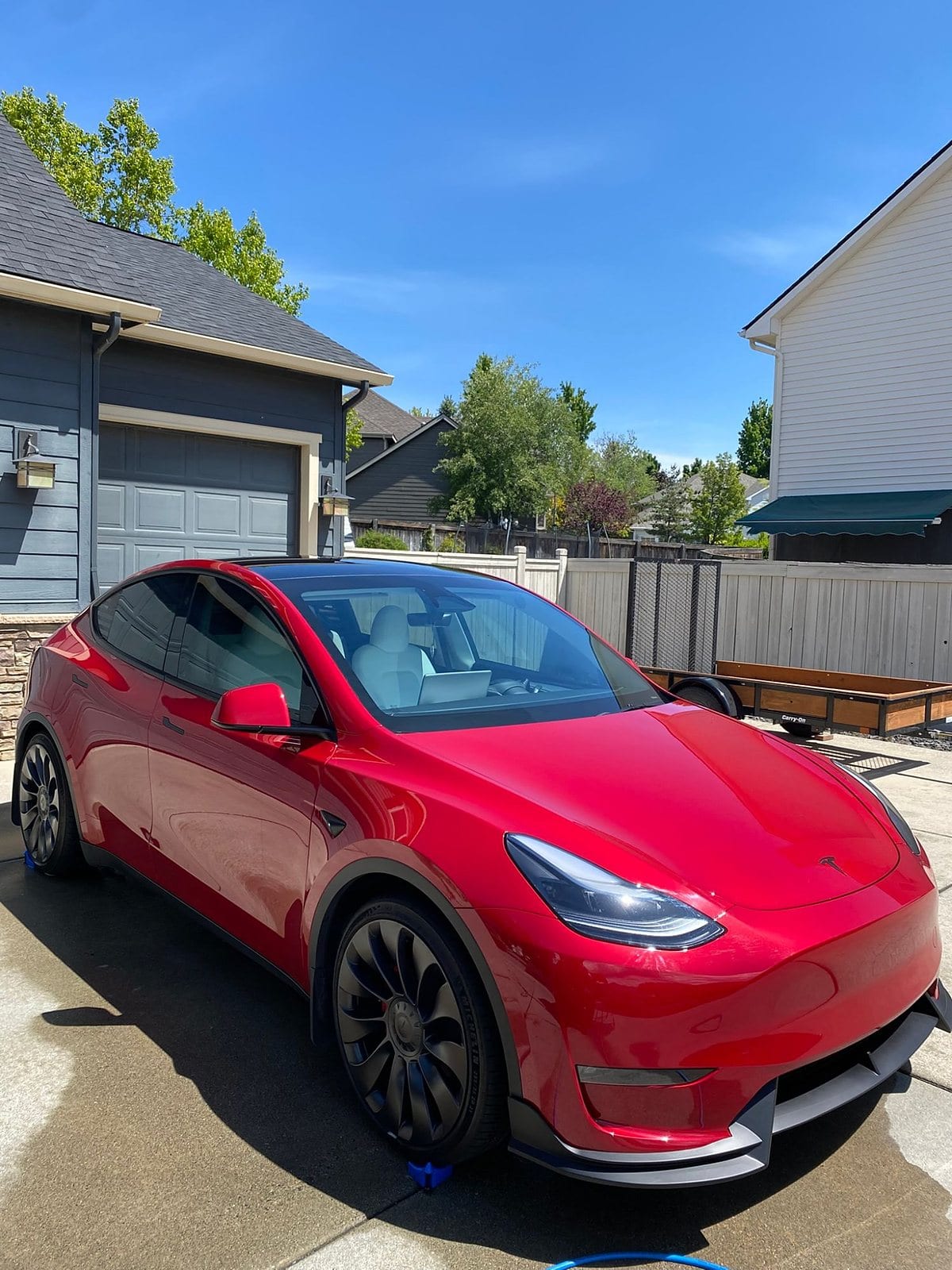 A red Tesla Model Y crossover with black wheels and a front splitter is parked in a driveway, showing a glossy, freshly detailed paint finish under bright sunlight.