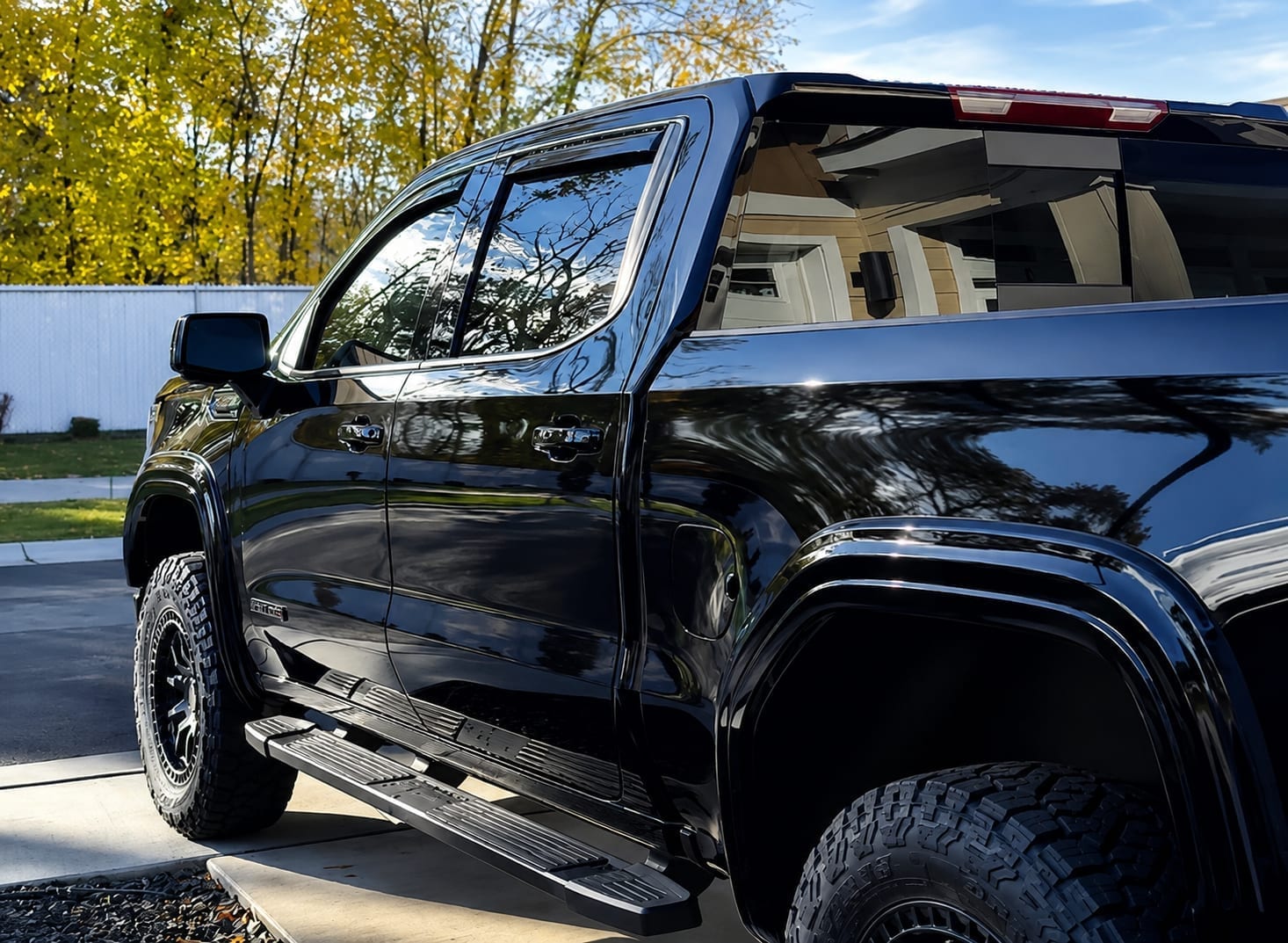 Close-up side profile of a black lifted pickup truck with off-road wheels and chrome trim, its glossy paint reflecting autumn trees, parked in a residential driveway.