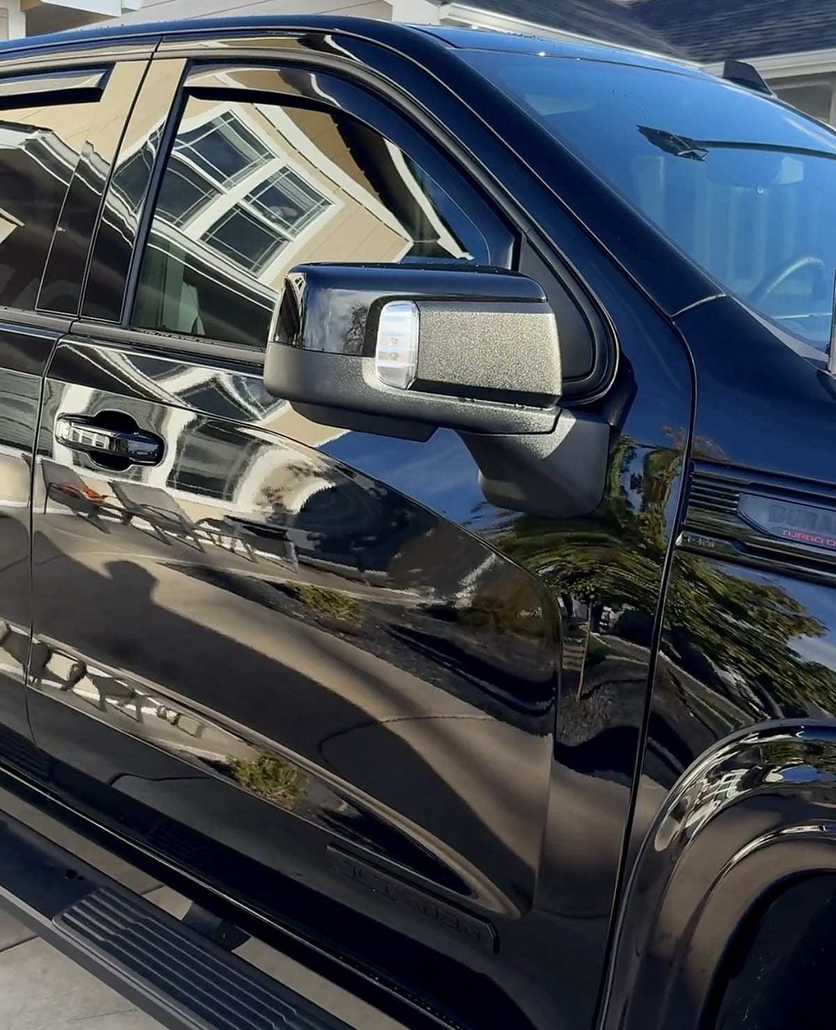 Close-up of a glossy black pickup truck’s side mirror and door panel, showing a deep, mirror-like reflective shine on the paint with a residential driveway reflected in the surface.