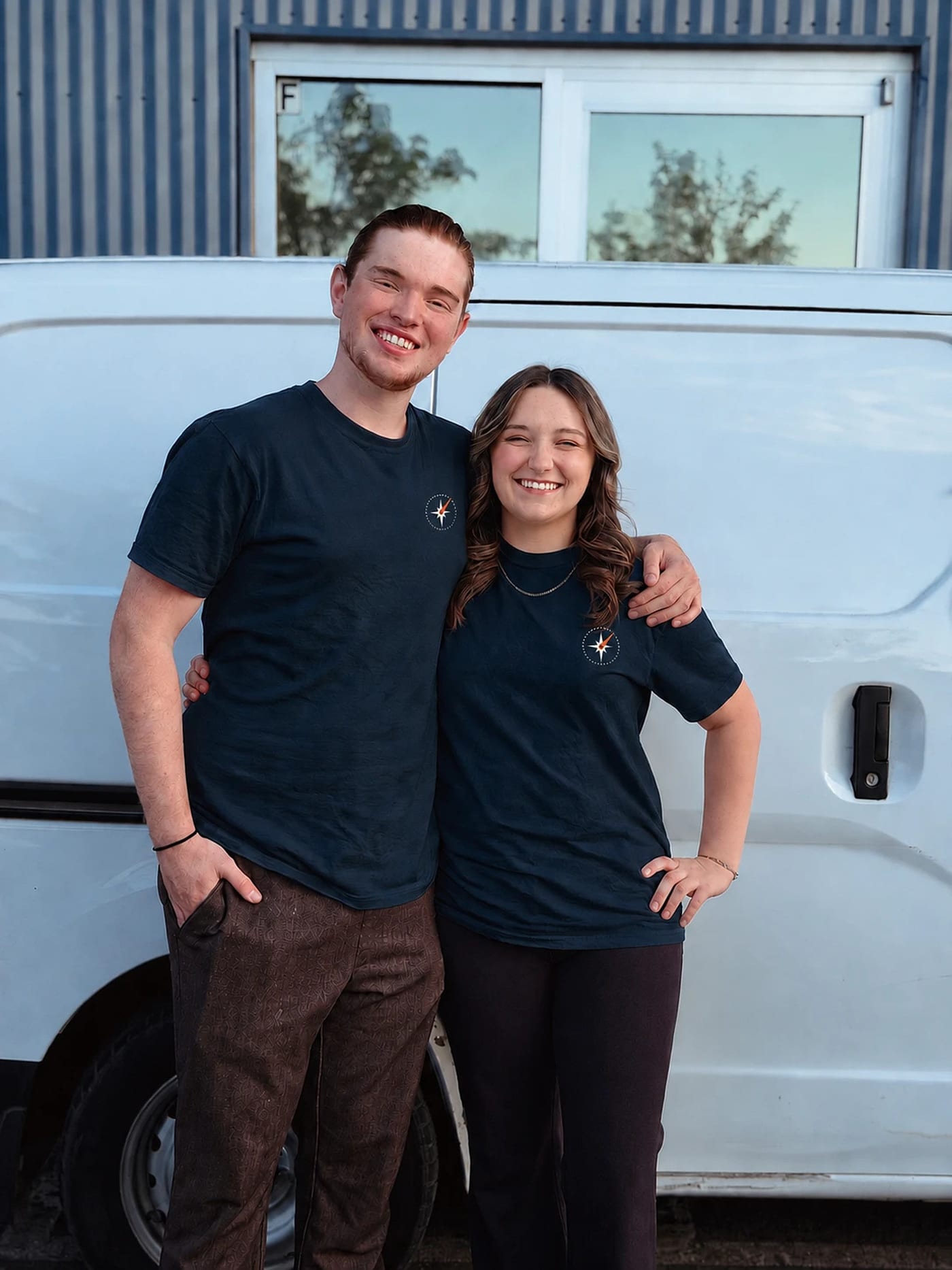 A man and a woman wearing matching navy Waypoint-branded t-shirts stand smiling in front of a white service van, with the man's arm around the woman's shoulder.