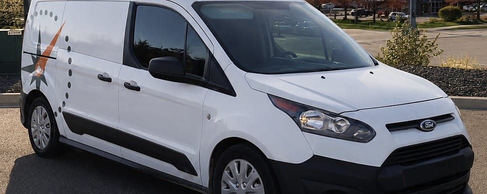 A white Ford Transit Connect cargo van with a partial compass-star logo decal parked in a parking lot, likely used as a mobile detailing service vehicle.